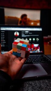 A hand holds a Rubik's Cube in front of a laptop with a blurred screen, indoors.