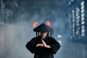 A martial artist in conical hat and black outfit performing a pose in an industrial setting.