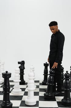 A young man in black contemplates his move on a large chessboard with oversized pieces.
