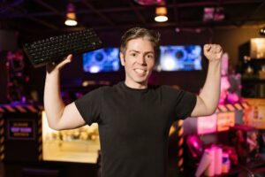 Smiling gamer in black shirt holding keyboard, celebrating indoors with gaming setup.